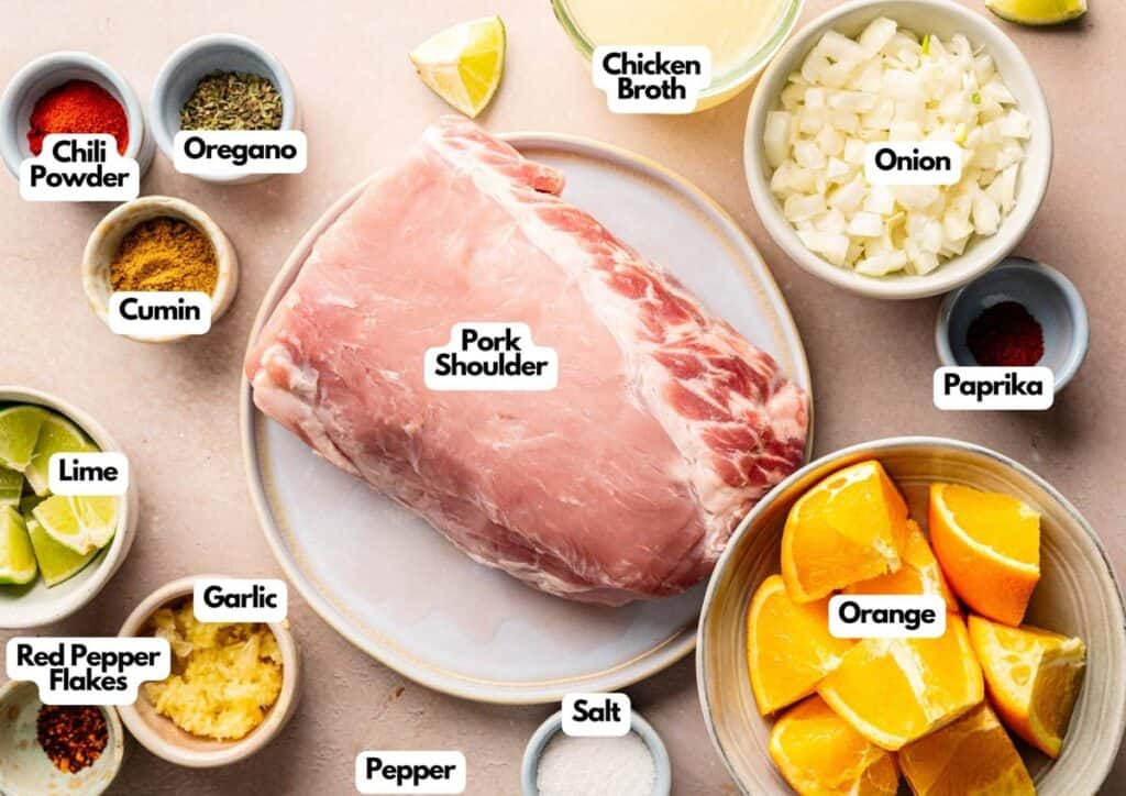 Raw pork shoulder surrounded by bowls of chopped onion, orange slices, garlic, lime wedges, chicken broth, and assorted spices&mdash;perfect ingredients for flavorful Slow Cooker Pork Carnitas.