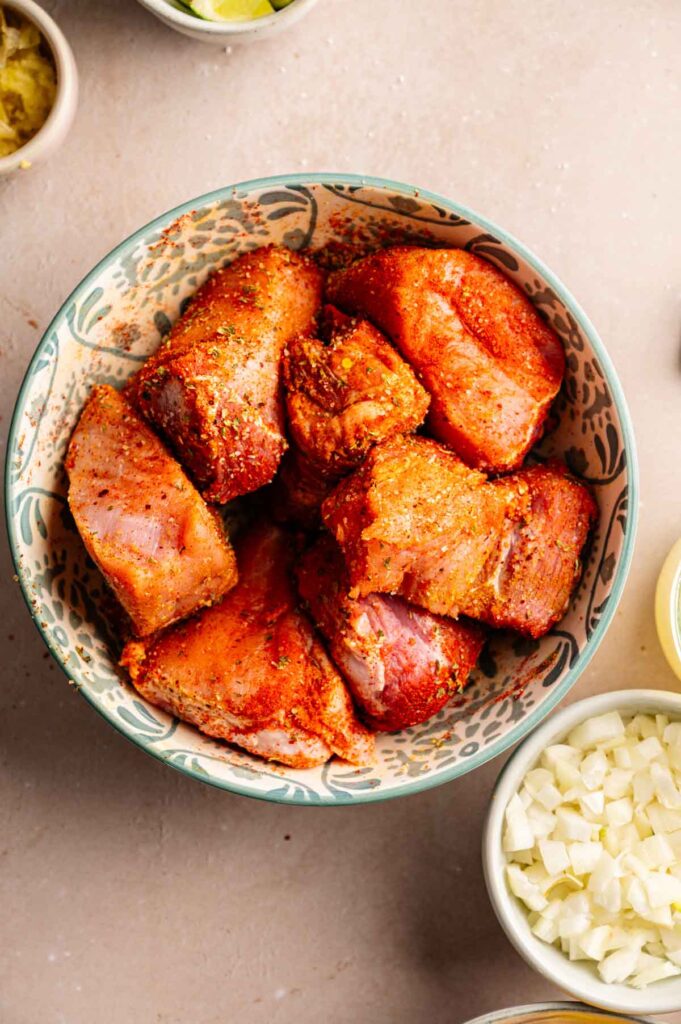 Raw pork shoulder pieces coated in a chili and cumin spice rub in a bowl.