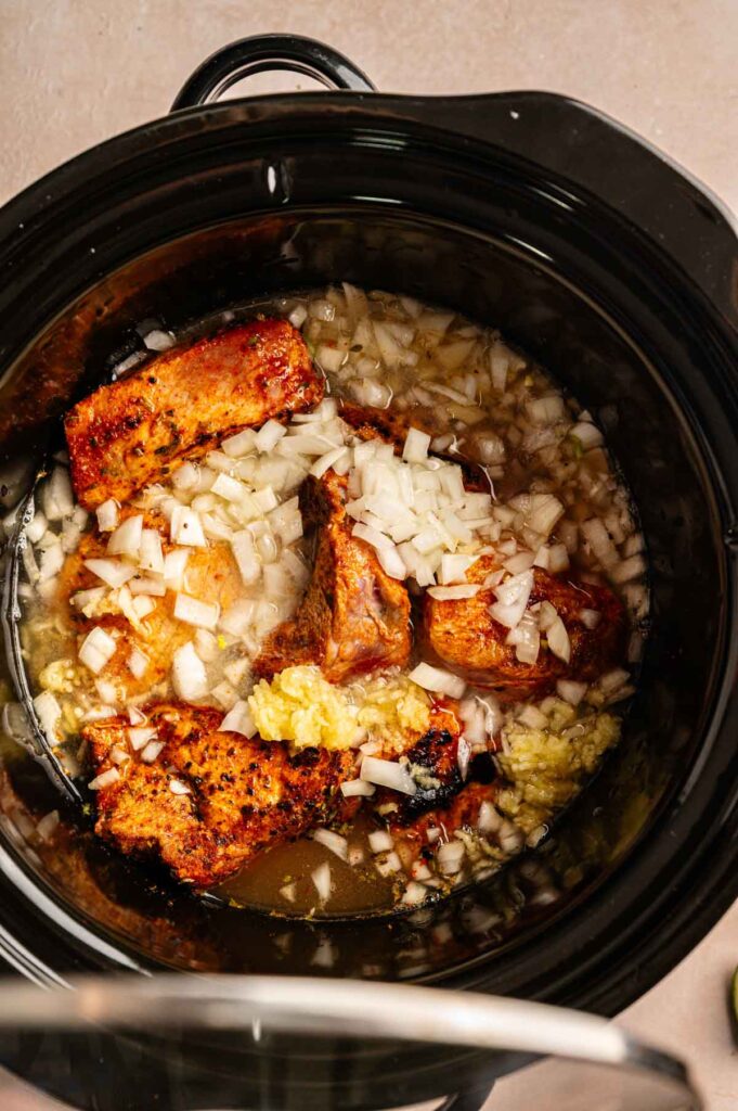 Seared pork pieces topped with diced onion and minced garlic in a slow cooker.