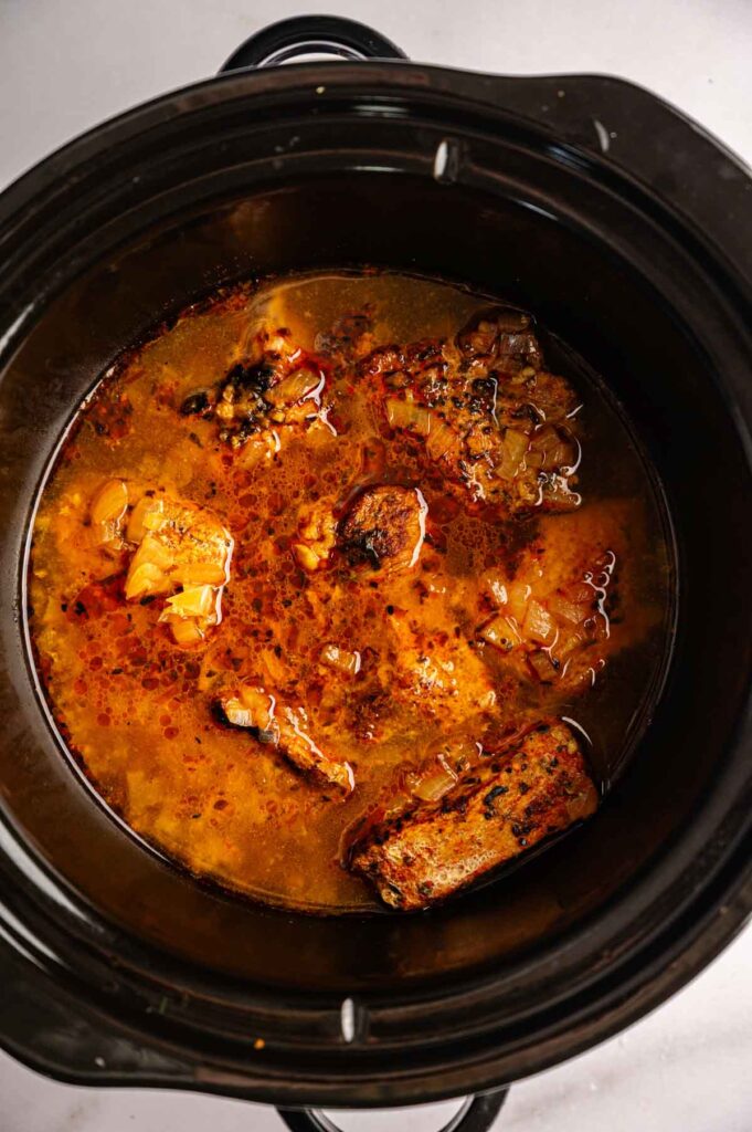 Chunks of meat in a reddish-brown broth with visible onions and spices, cooking inside a black slow cooker—perfect for Slow Cooker Pork Carnitas.
