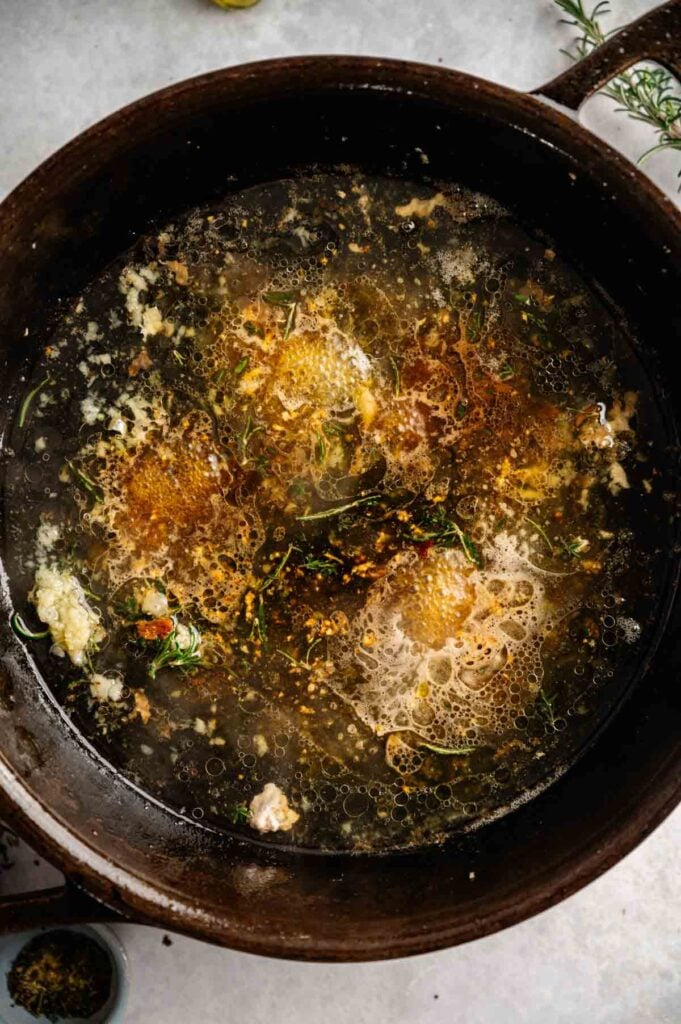 Chicken broth simmering in a skillet with garlic and rosemary, scraping up browned bits.