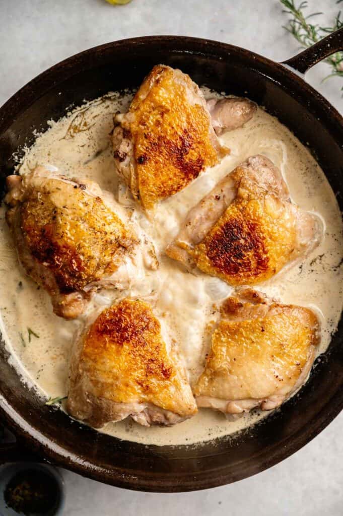 Five browned Tuscan Chicken Thighs simmer in a creamy white sauce inside a black skillet.