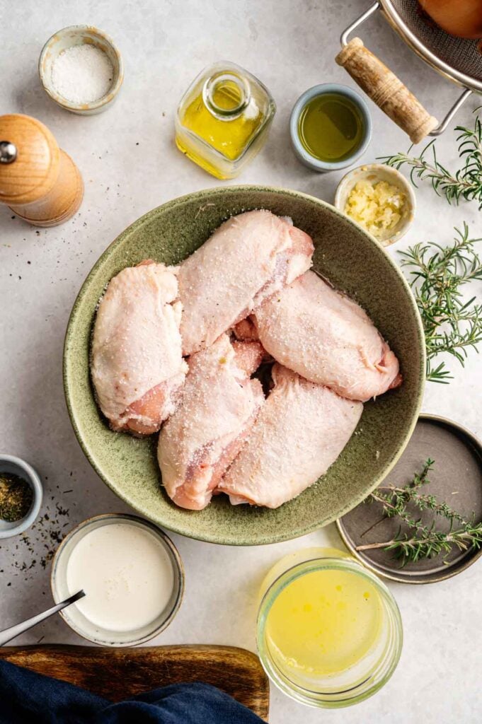 Raw, seasoned chicken thighs in a bowl surrounded by olive oil, garlic, and fresh rosemary.