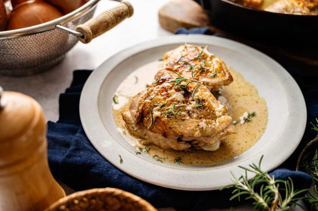Two pieces of Tuscan Chicken Thighs with golden crispy skin served on a white plate, topped with creamy sauce and herbs, surrounded by kitchen items.