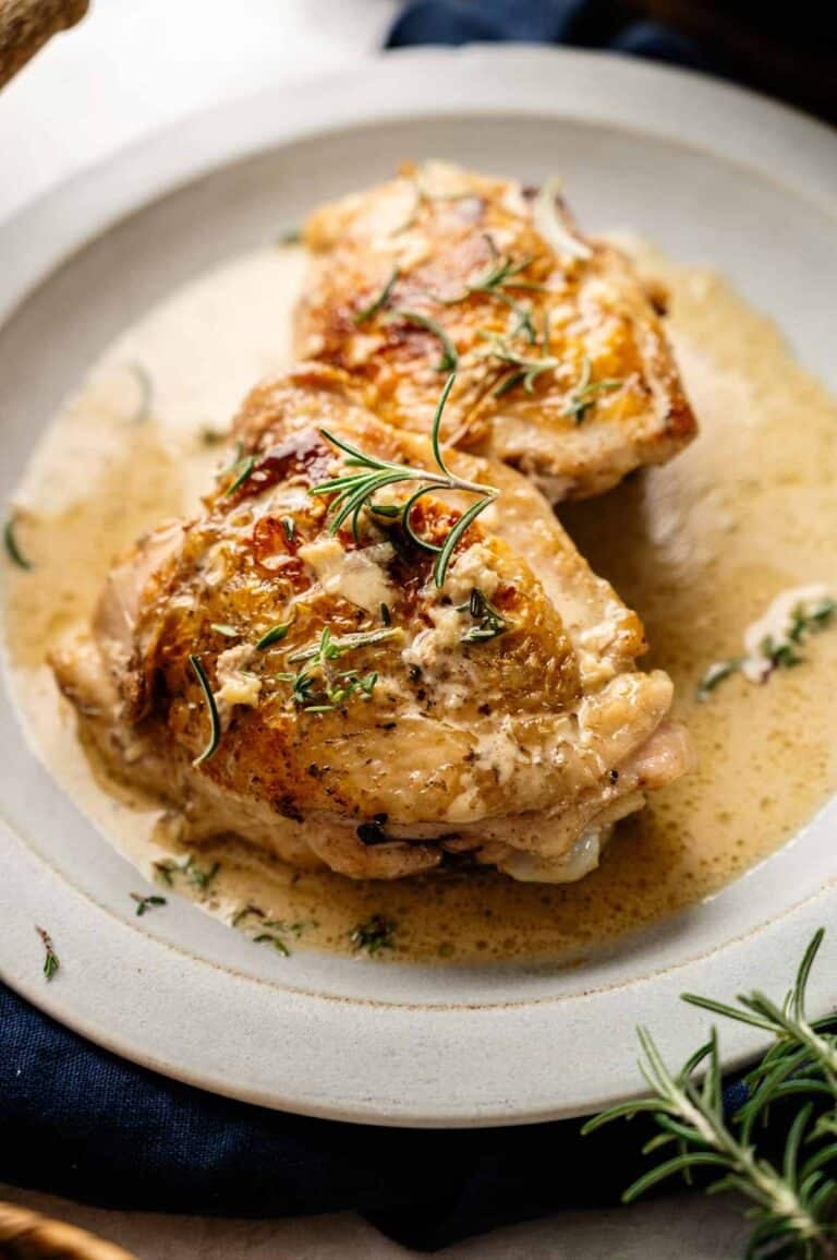Two pieces of roasted Tuscan Chicken Thighs with crispy skin are served on a plate with creamy sauce and garnished with fresh rosemary.