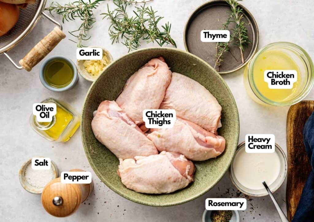 A bowl of raw Tuscan Chicken Thighs is surrounded by labeled ingredients: garlic, thyme, chicken broth, olive oil, heavy cream, rosemary, salt, and pepper.