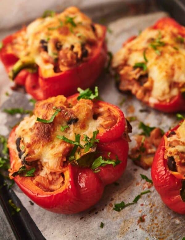 Four baked red bell pepper halves stuffed with rice, vegetables, and melted cheese, garnished with chopped herbs, on a parchment-lined baking tray.