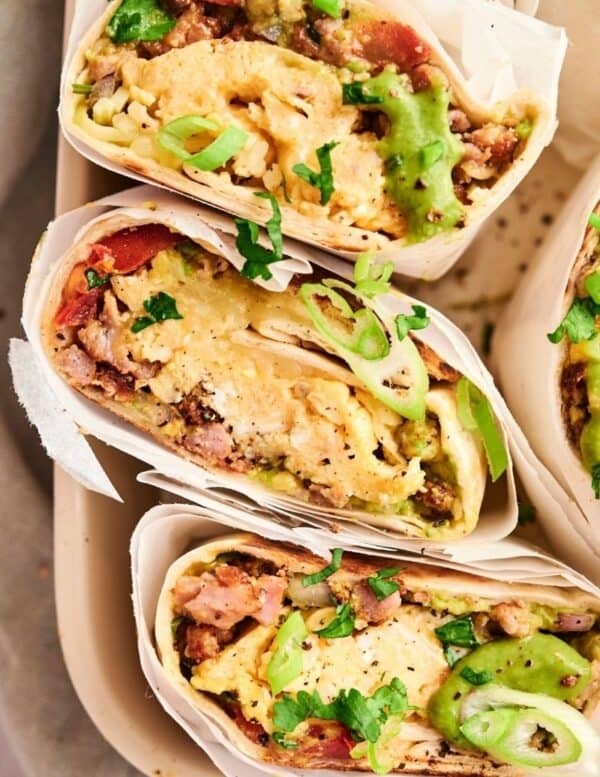 Three breakfast burritos cut in half, filled with scrambled eggs, sausage, avocado, and topped with sliced green onions and cilantro.