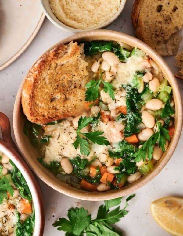 A bowl of vegetable and white bean soup topped with parsley and Parmesan, served with a slice of toasted bread, with lemon, herbs, and more bread nearby.