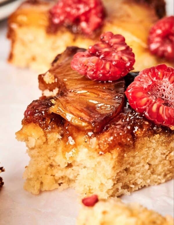 A close-up of a slice of pineapple upside-down cake topped with caramelized pineapple and fresh raspberries.