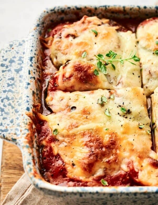 A close-up of a baked lasagna in a speckled ceramic dish, topped with melted cheese and garnished with fresh herbs.