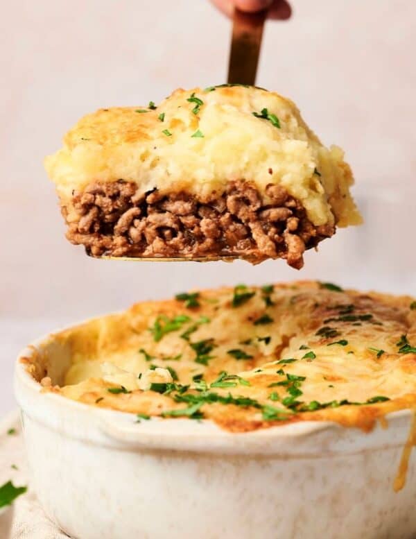 A serving of shepherd’s pie with a layer of ground meat topped with mashed potatoes is lifted from a baking dish, garnished with chopped herbs.