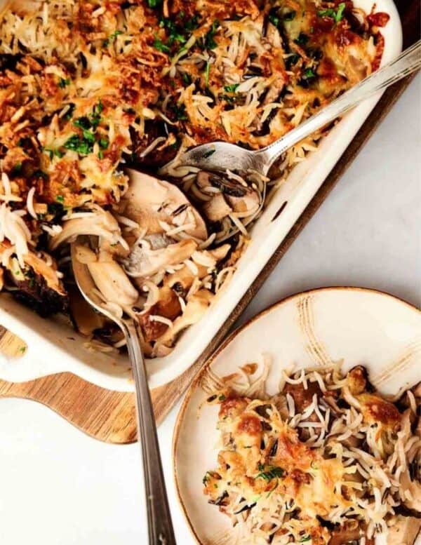 A baked rice and mushroom casserole in a dish with a spoon, served onto a plate beside it.