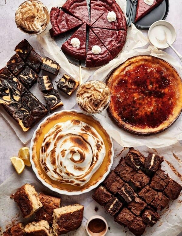 An assortment of desserts including pies, brownies, cake slices, topped with whipped cream and caramelized or toasted finishes, arranged on a table.