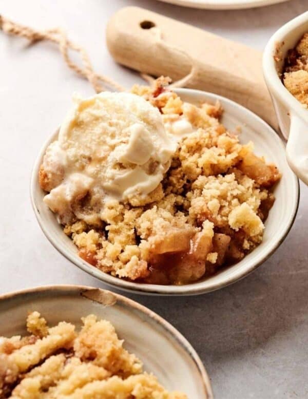 A bowl of apple crumble topped with a scoop of vanilla ice cream, with a wooden utensil and another dish of crumble in the background.