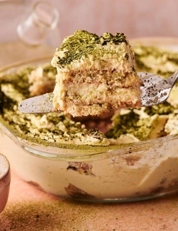 A slice of layered tiramisu, topped with green matcha powder, is being lifted from a glass dish with a spatula.