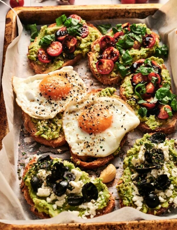 A tray of avocado toast topped with fried eggs, sliced cherry tomatoes, fresh herbs, black olives, and crumbled cheese on parchment paper.