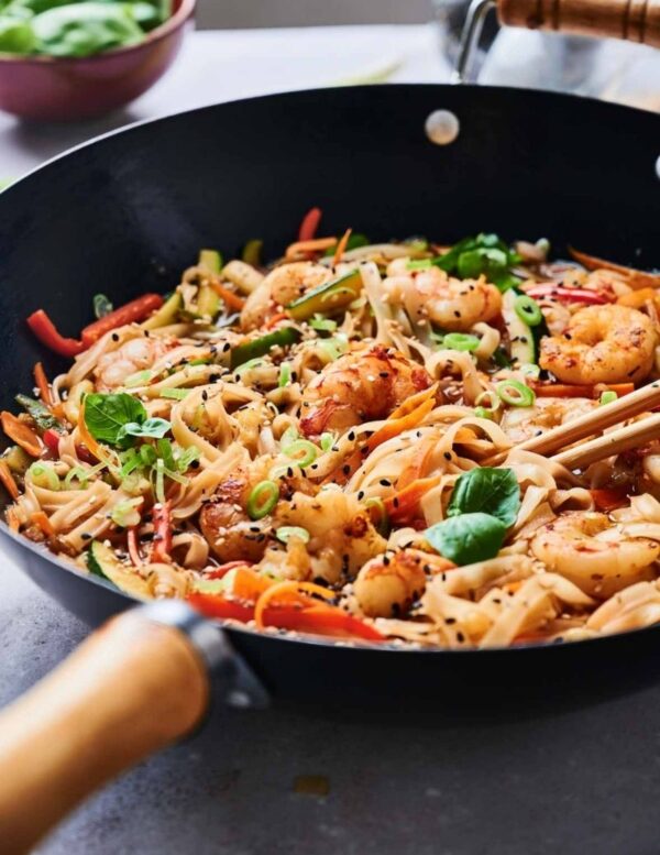 A wok filled with shrimp stir-fry, noodles, sliced vegetables, green onions, black sesame seeds, and fresh basil leaves.