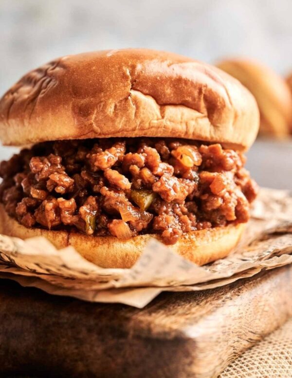A sloppy joe sandwich filled with seasoned ground meat and sauce, served on a hamburger bun and placed on brown parchment paper.