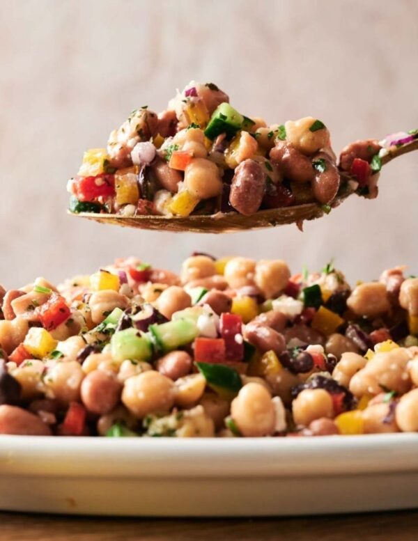 A close-up of a spoonful of mixed bean salad with chopped vegetables, held above a plate filled with more of the same salad.