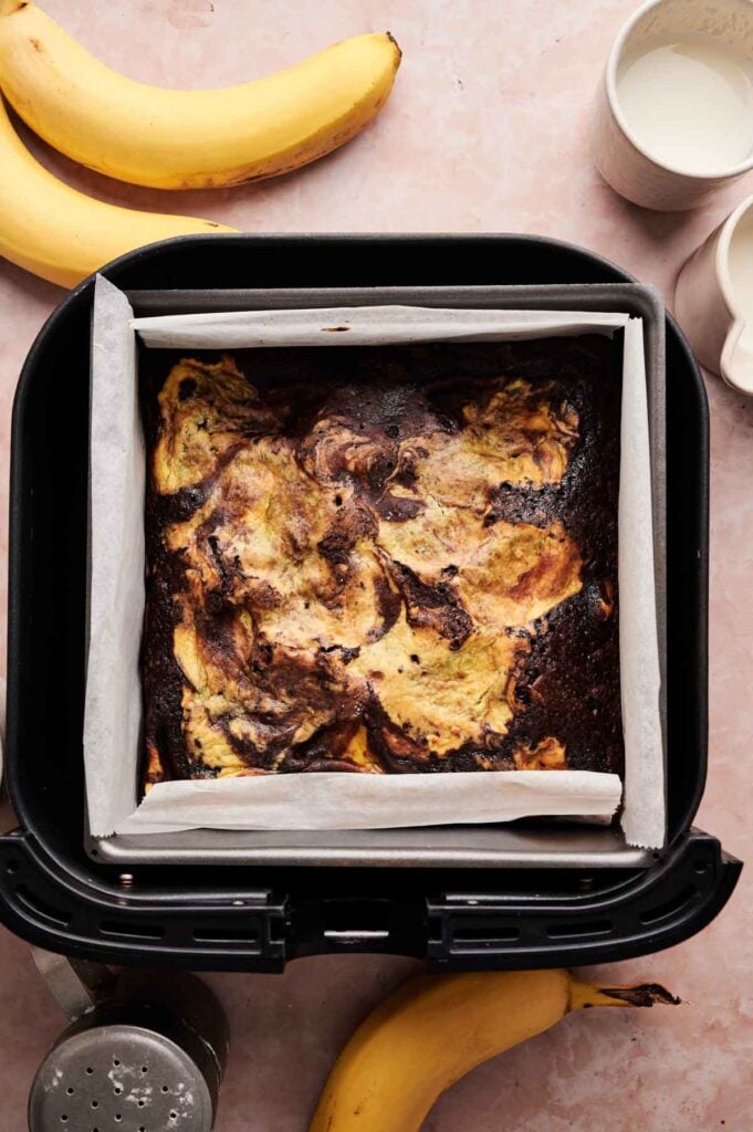 A batch of Air Fryer Banana Pudding Brownies rests in a square parchment-lined pan inside an air fryer, surrounded by bananas and small pitchers of milk on a pink surface.