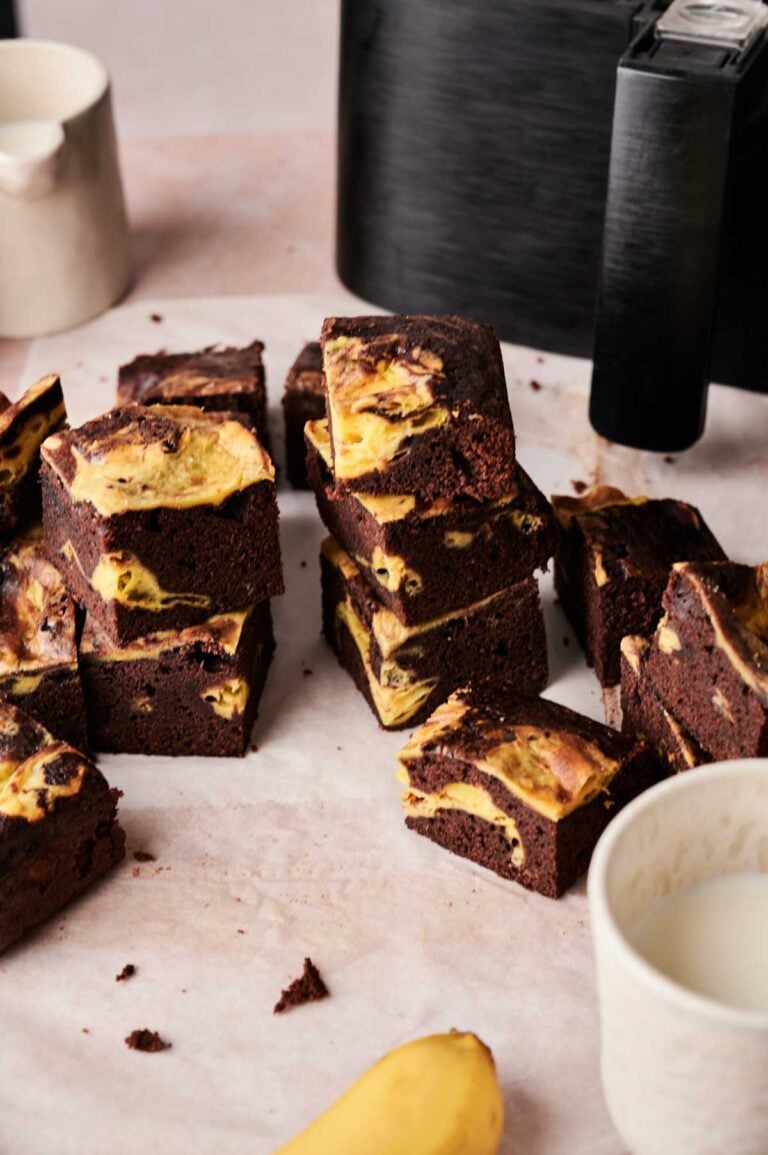 Air Fryer Banana Pudding Brownies with a marbled yellow swirl are stacked on a light surface, with a banana, a cup, and a black appliance in the background.