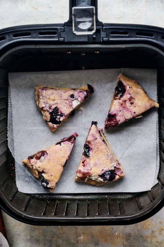 Four unbaked triangular Air Fryer Blueberry Scones rest on parchment paper inside an air fryer basket, ready to bake.