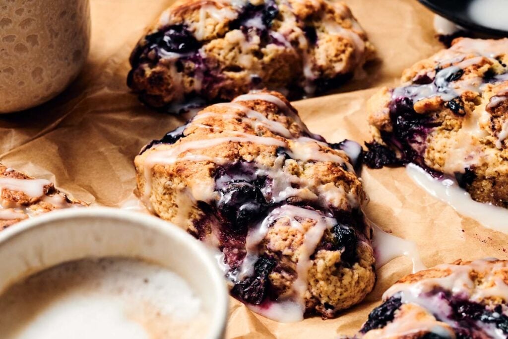 Close-up of Air Fryer Blueberry Scones drizzled with icing on parchment paper, with a partially visible cup of coffee in the foreground.