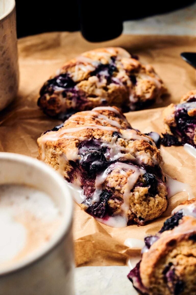 Close-up of Air Fryer Blueberry Scones with icing drizzled on top, placed on brown parchment paper, with a cup of coffee in the foreground.