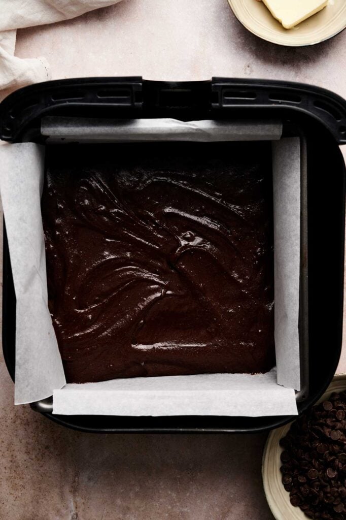 A square pan lined with parchment paper is filled with brownie batter, ready for baking. Butter and chocolate chips are visible nearby.