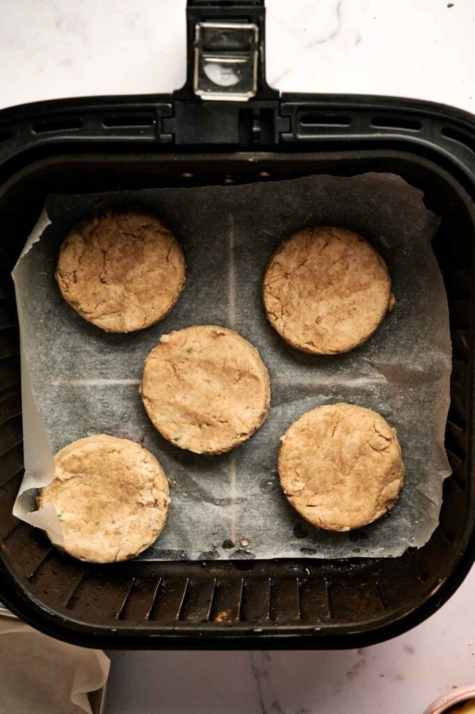 Five round, uncooked Air Fryer Cheddar Biscuits are arranged on parchment paper inside an air fryer basket, ready to be baked.