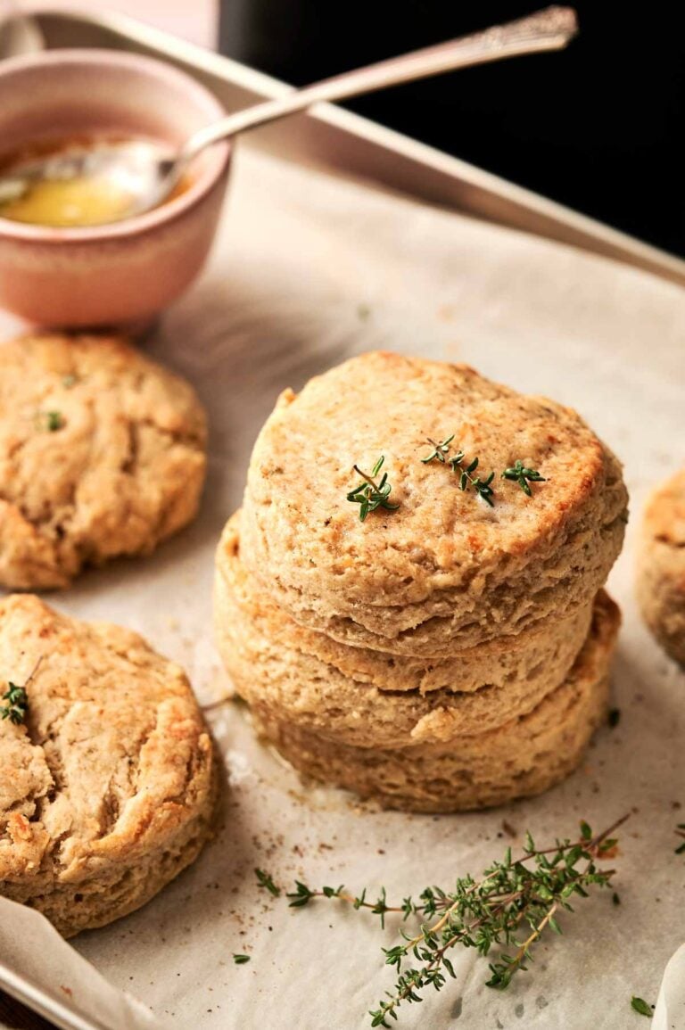 Three stacked Air Fryer Cheddar Biscuits on parchment paper, garnished with fresh thyme, surrounded by more biscuits and a small bowl of melted butter in the background.