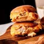 Two Air Fryer Cheeseburger Sliders with melted cheese are stacked on a wooden board, with a blurred background and a beige cloth nearby.