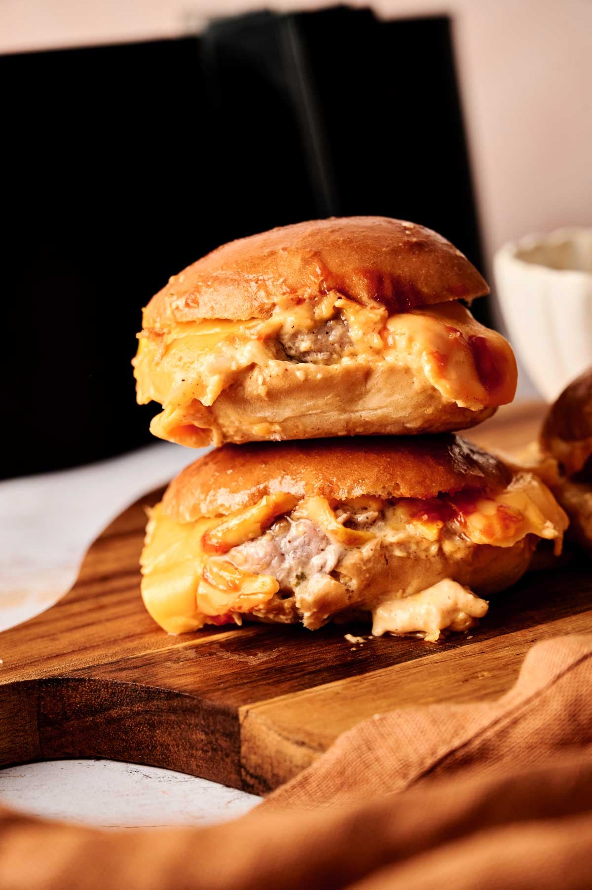 Two Air Fryer Cheeseburger Sliders with melted cheese are stacked on a wooden board, with a blurred background and a beige cloth nearby.