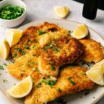 A plate of crispy Air Fryer Chicken Cutlets garnished with chopped herbs and lemon wedges, with a small bowl of herbs and a bottle of oil in the background.