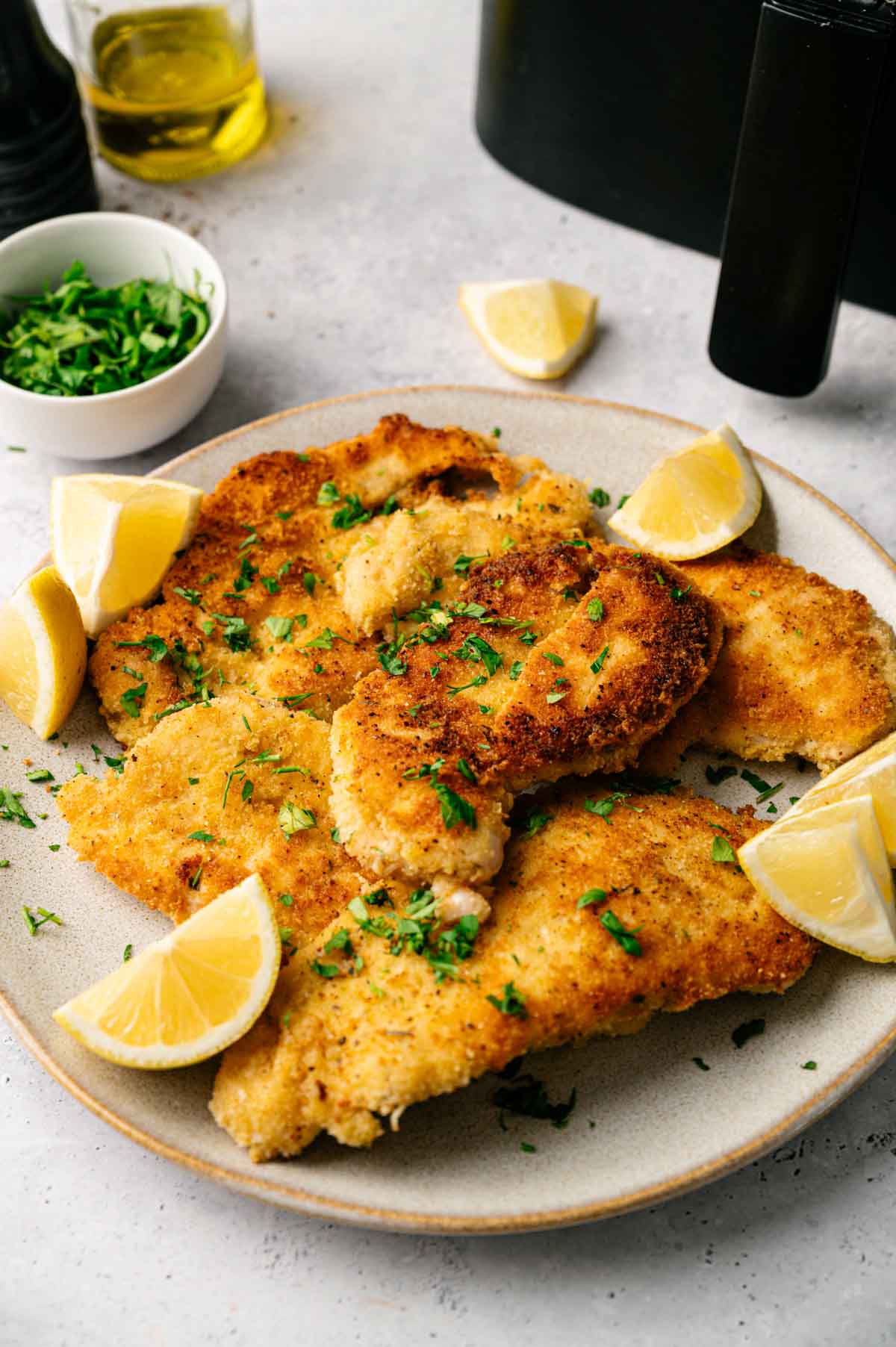 A plate of crispy Air Fryer Chicken Cutlets garnished with chopped herbs and lemon wedges, with a small bowl of herbs and a bottle of oil in the background.