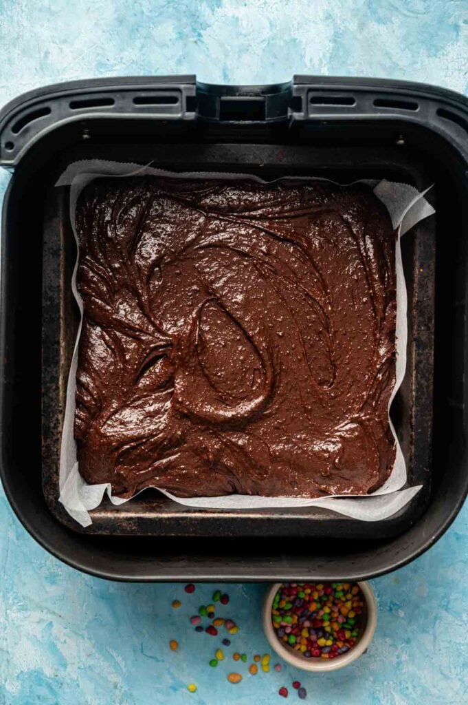 Brownie batter spread in a parchment-lined tray inside an air fryer basket, ready to become delicious Air Fryer Cosmic Brownies, with a small bowl of colorful candy pieces nearby.