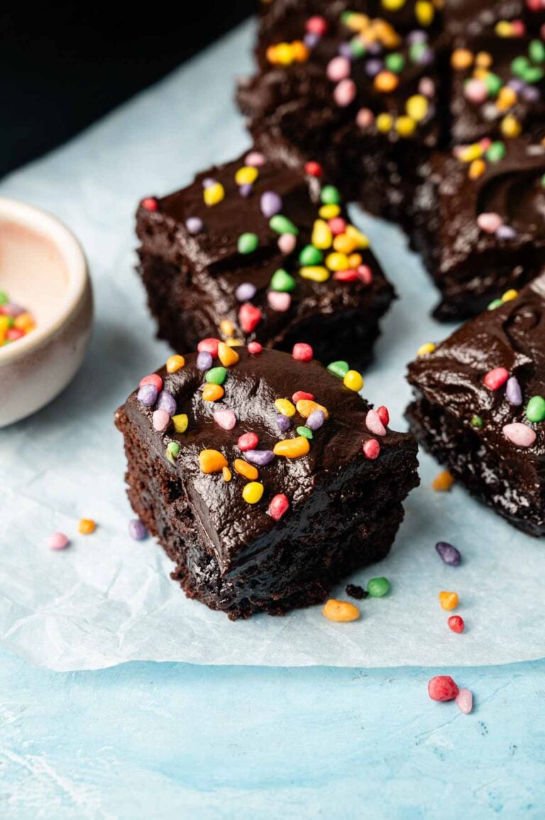 Several pieces of chocolate frosted Air Fryer Cosmic Brownies topped with colorful round sprinkles are arranged on a sheet of parchment paper.