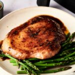 A cooked pork chop with sauce rests on a bed of steamed asparagus spears, served on a beige plate—perfect alongside quick Air Fryer Ham Steaks for an easy, flavorful meal.