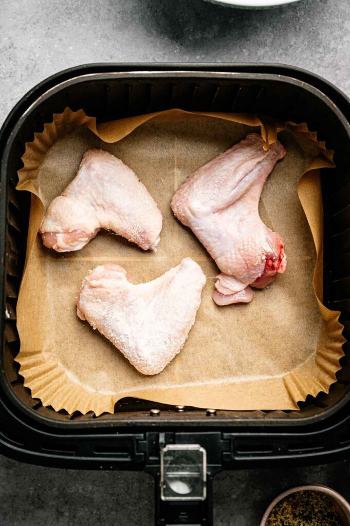 Three raw chicken wings placed on parchment paper in an air fryer basket, ready to be transformed into flavorful Air Fryer Lemon Pepper Wings.
