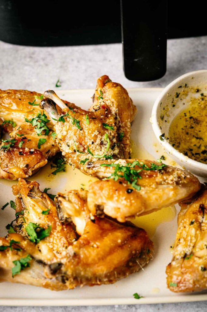 Five cooked chicken wings garnished with chopped herbs are arranged on a white tray next to a small bowl of dipping sauce—these Air Fryer Lemon Pepper Wings offer the perfect balance of crispy texture and zesty flavor.