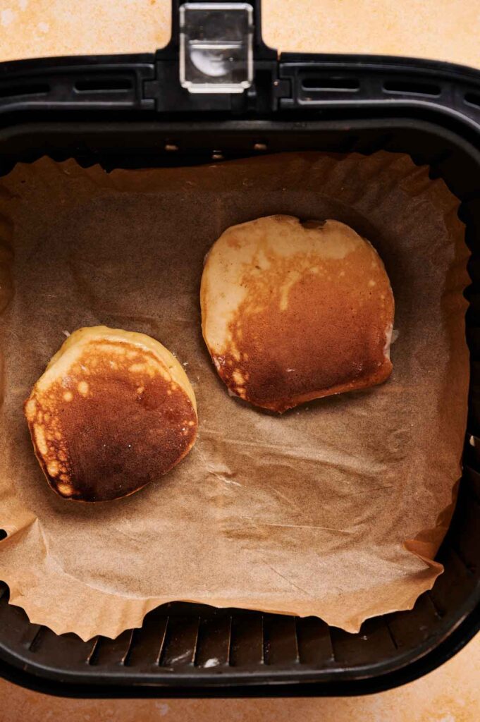 Two Air Fryer Lemon Ricotta Pancakes with browned tops sit on parchment paper inside an air fryer basket.