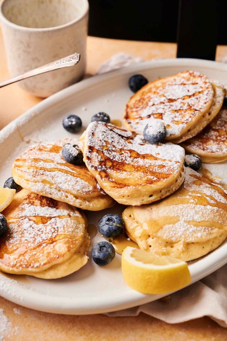 A plate of fluffy Air Fryer Lemon Ricotta Pancakes topped with powdered sugar, fresh blueberries, and lemon wedges, with a cup and a fork in the background.