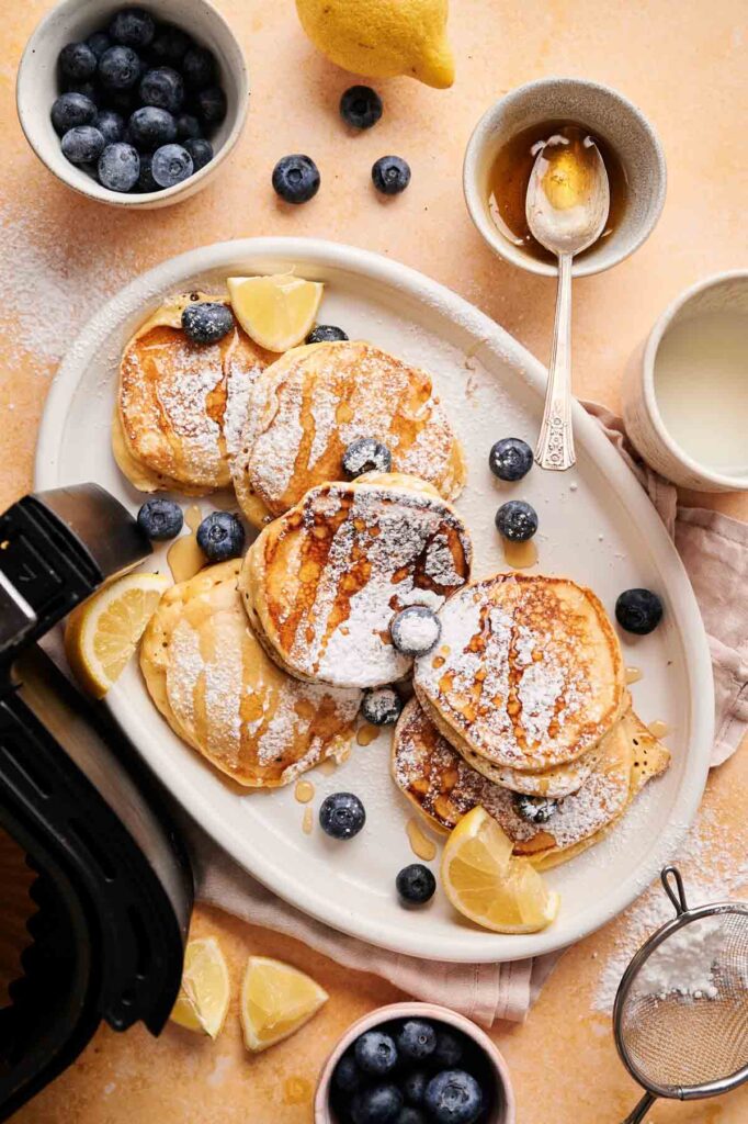 A plate of Air Fryer Lemon Ricotta Pancakes dusted with powdered sugar, topped with blueberries and lemon wedges, surrounded by a bowl of blueberries, honey, a lemon, and a cup of milk.