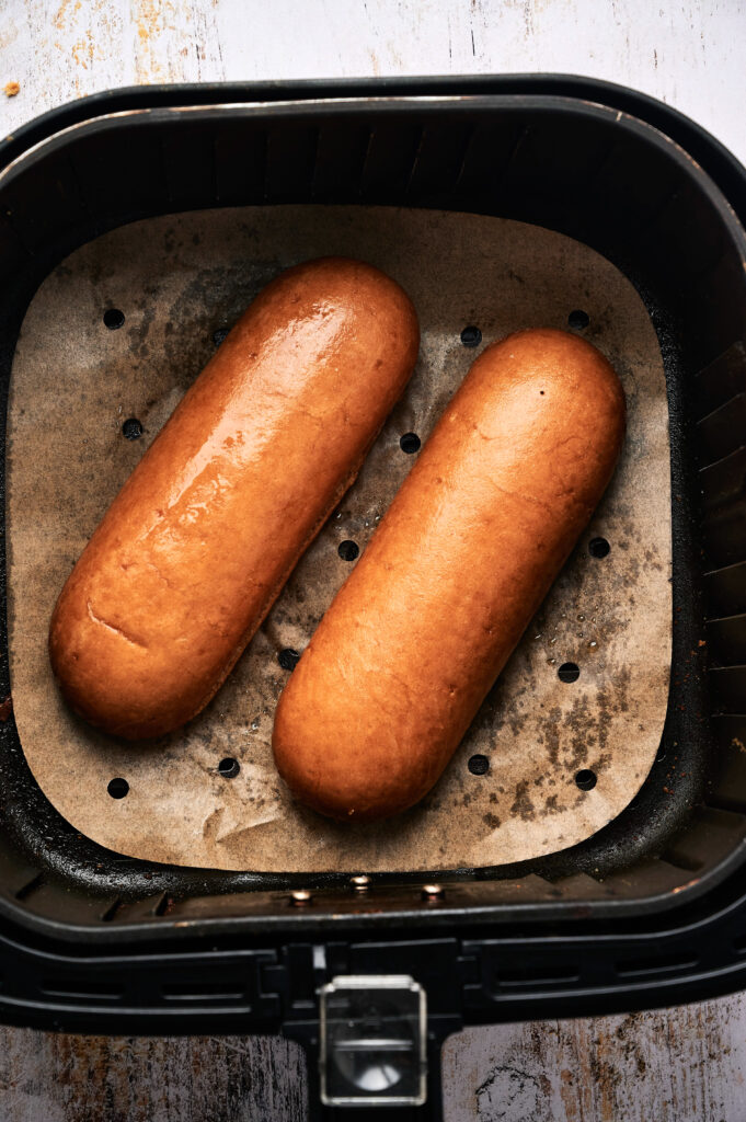 Two golden brown bread rolls on parchment paper inside an air fryer basket, perfect for crafting a delicious Air Fryer Lobster Roll.