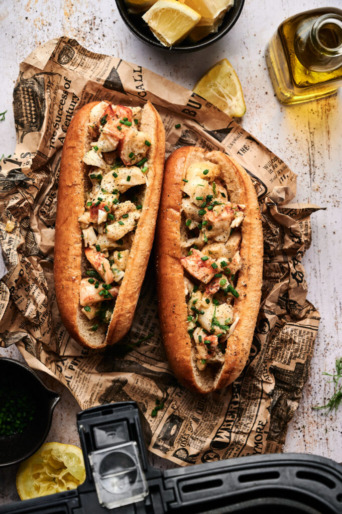 Two Air Fryer Lobster Rolls filled with lobster meat and herbs on buns, placed on newspaper-style paper. Lemon wedges, olive oil, and chopped herbs are nearby for the perfect finishing touch.