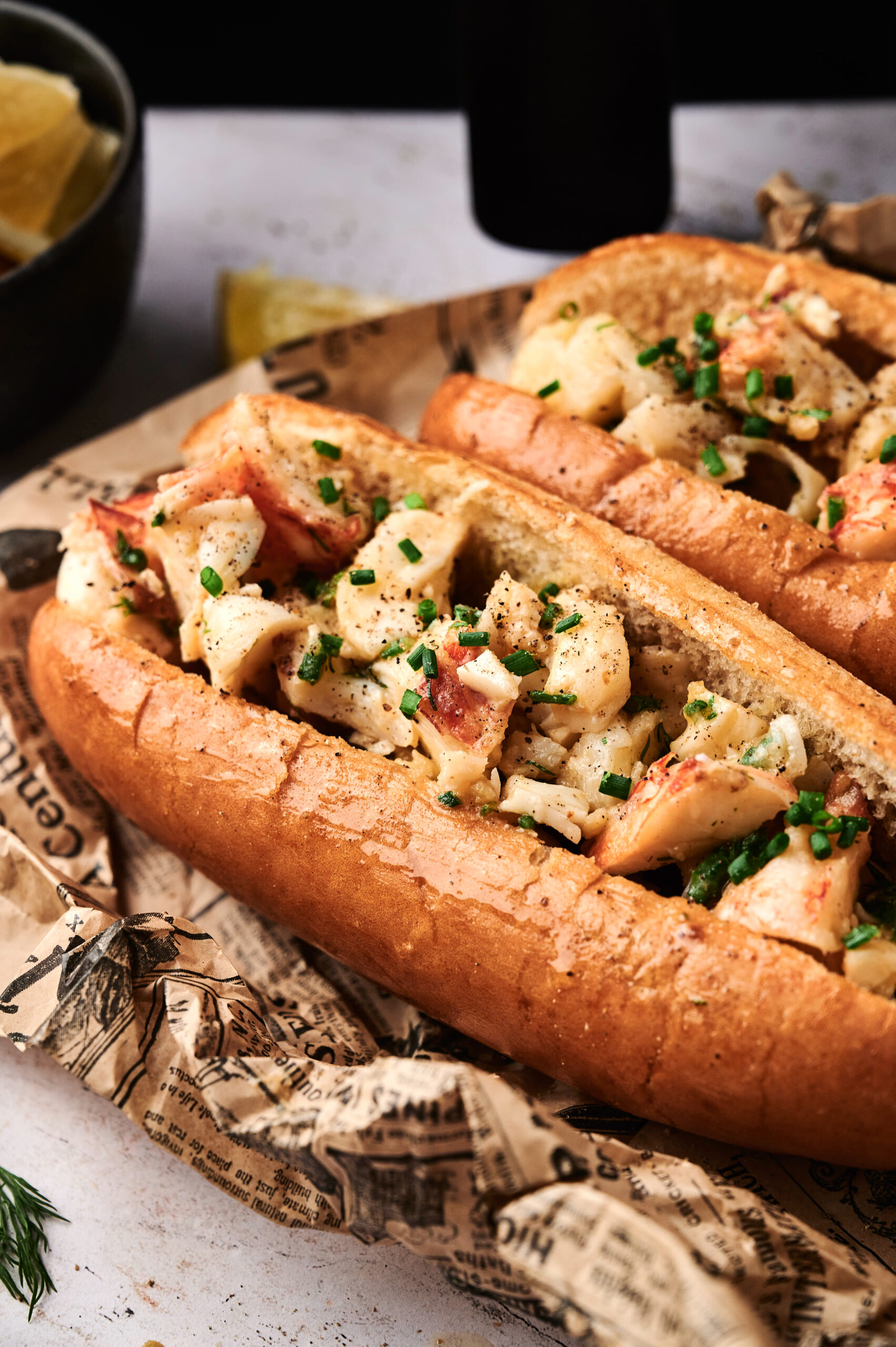 A close-up of an Air Fryer Lobster Roll with chunks of lobster meat in a toasted bun, garnished with chopped chives, on newspaper-style paper.