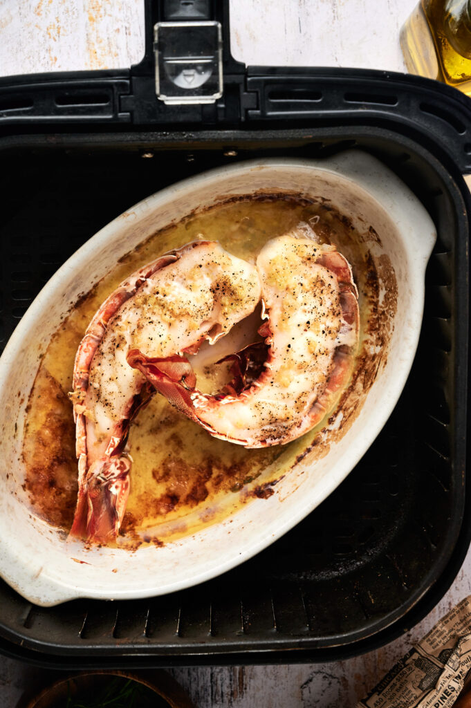 Two seasoned lobster tails are cooked in a white oval dish inside an air fryer, perfect for creating a flavorful Air Fryer Lobster Roll.