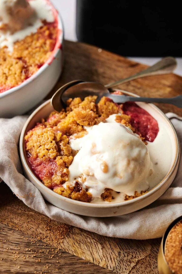 A bowl of Air Fryer Rhubarb Crisp topped with a scoop of vanilla ice cream, with a spoon on the side and a baking dish in the background.
