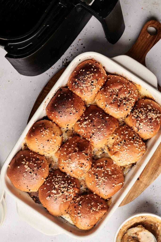 A baking dish filled with twelve golden-brown Air Fryer Roast Beef Sliders topped with seeds, next to an air fryer on a light countertop.